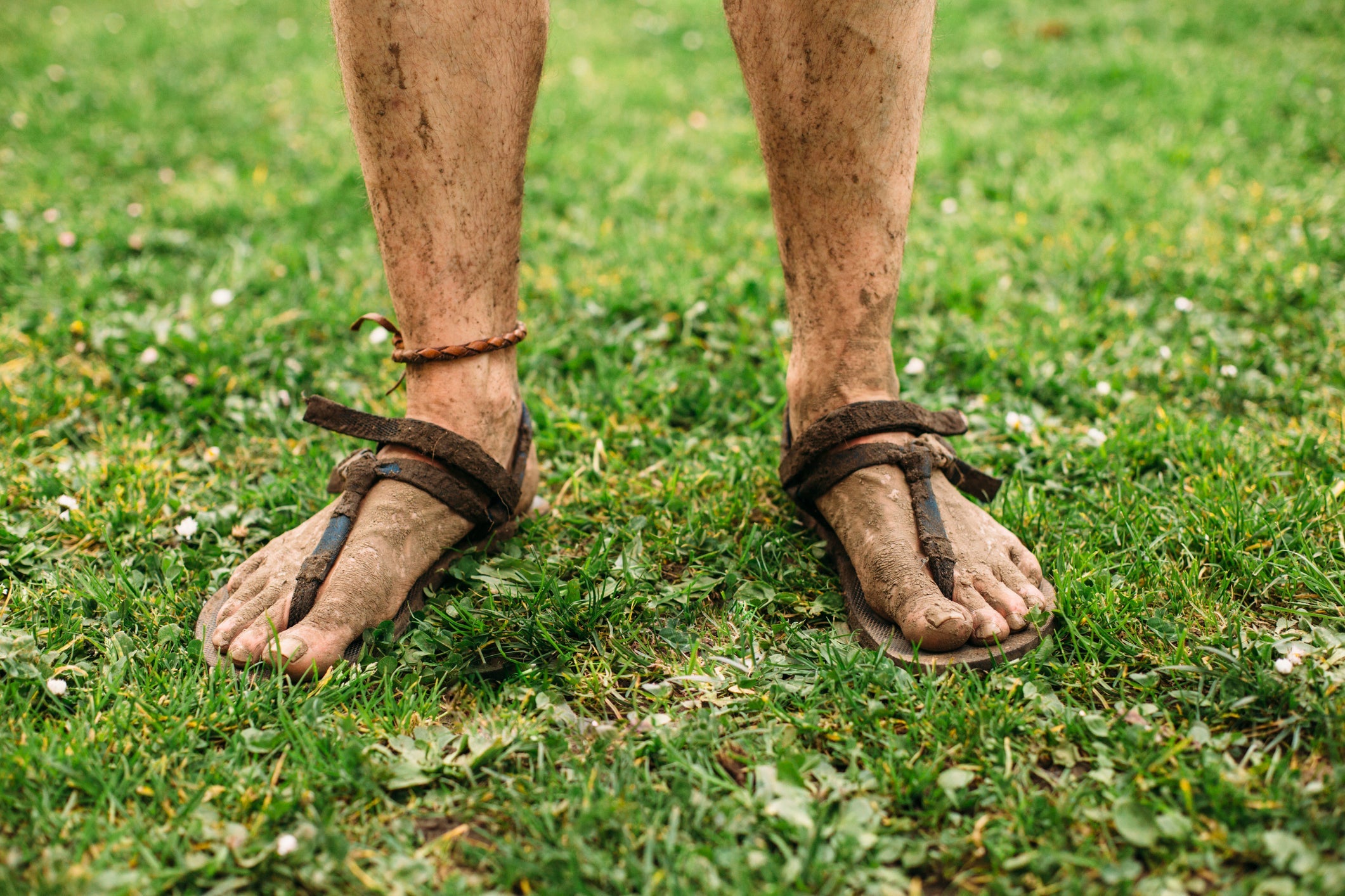 Mud stained minimalist runner feet and legs.
The runner is standing in green grass wearing barefoot sandals and a bracelet in their ankle.