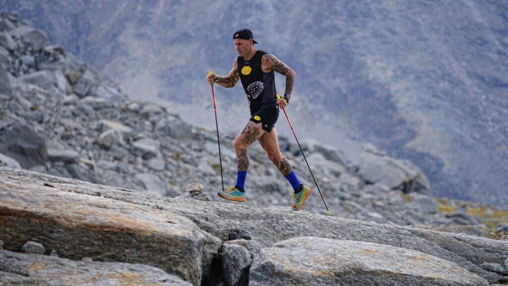 A man runs up a slab of rocks with trekking poles and backward hat