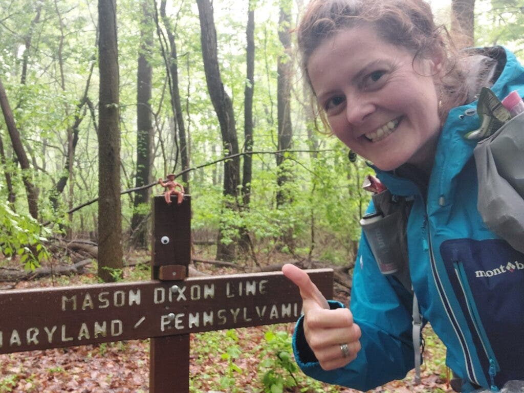 Heather Anderson smiles with a thumbs up in front of a Mason Dixon line sign on the trail. 