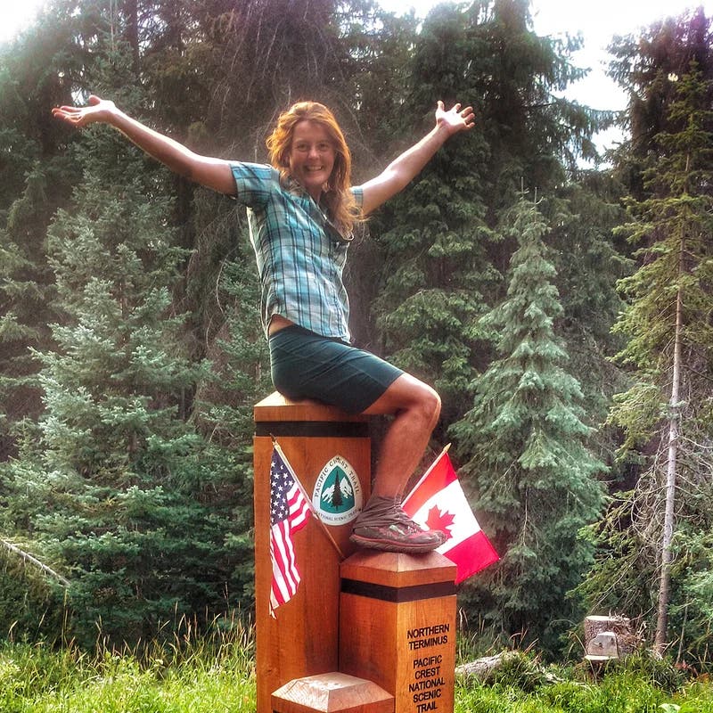 Heather Anderson sits with her arms raised on the marker designating the northern terminus of the PCT.
