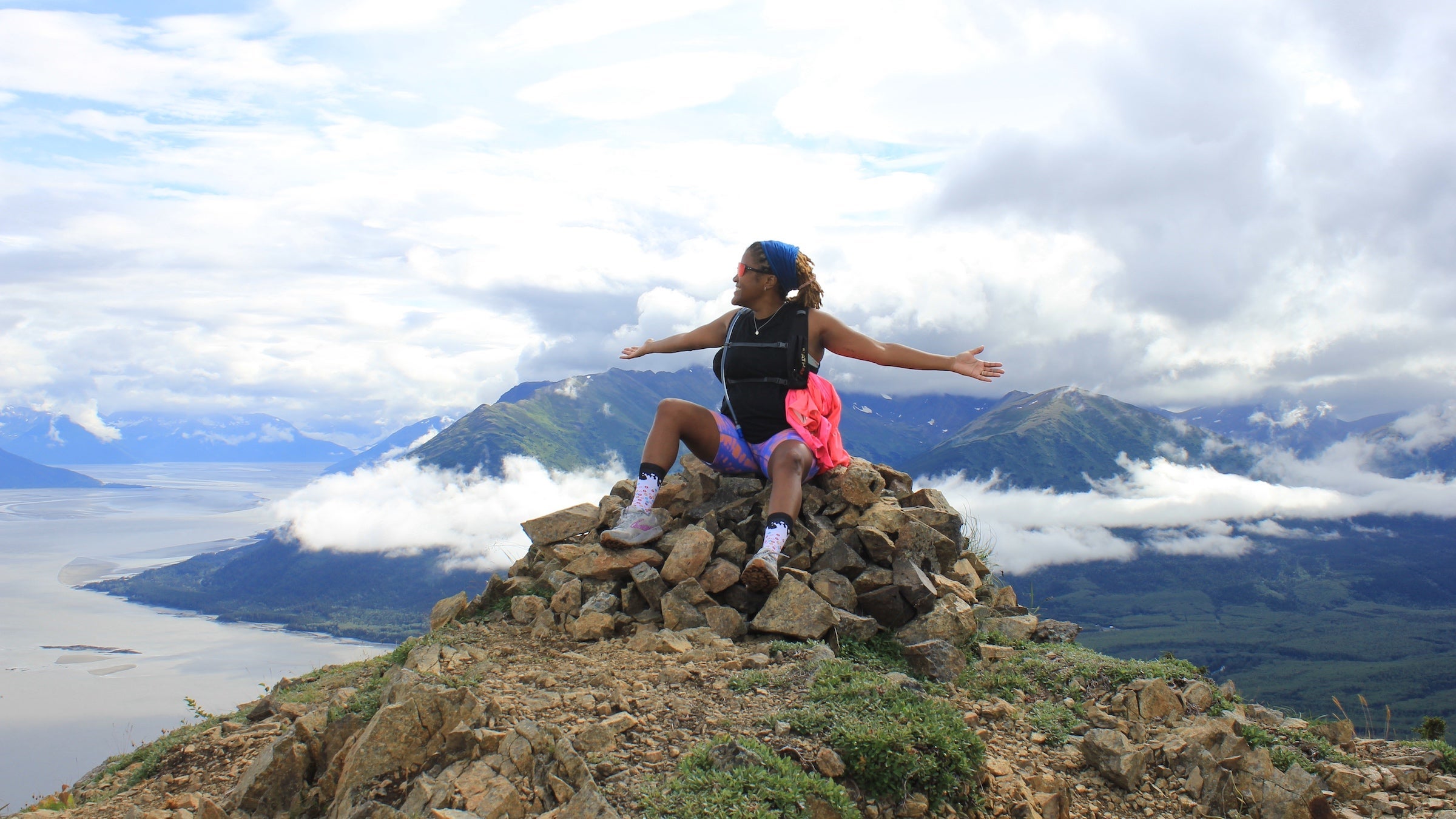 A woman in pink running bottoms spreads out her arms at the top of a mountain