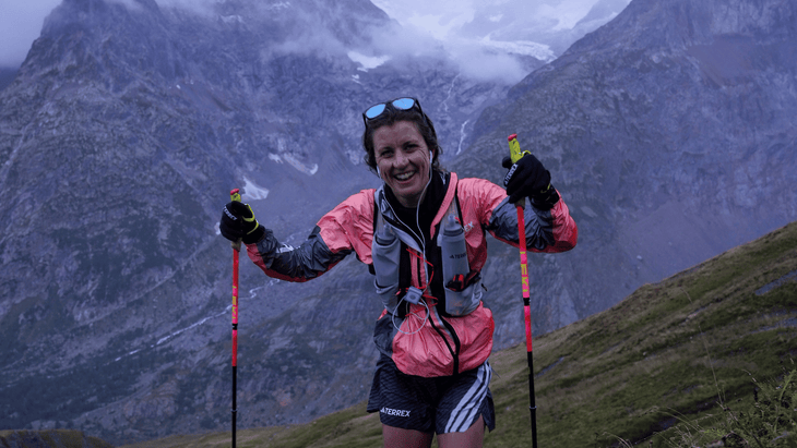 Ruth Croft in full cold weather kit using poles to climb an alpine pass with a glacier in the background.