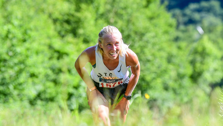 blond woman in running clothes is bent over as she runs up steep trail with green trees in background