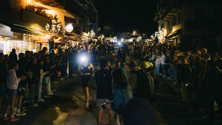 Photo of runners illuminated by headlamps making their way through a crowd in an alpine village.