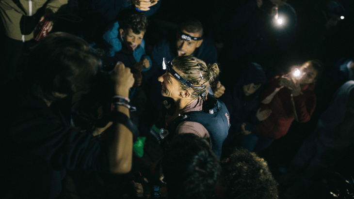Photograph of Courtney Dauwalter making her way through an aid station at night, illuminated by headlamps.