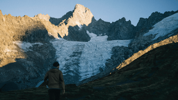 Photo of a glaciated alpine peak with a hiker in the foreground walking across a muddy trail.